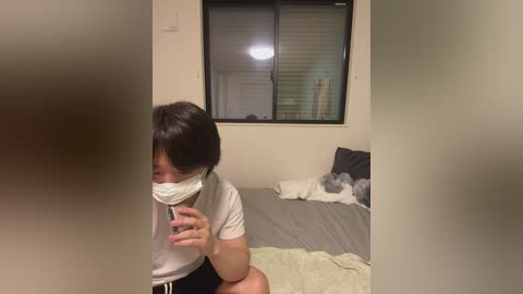 Video of a young Asian woman with short black hair and a white face mask, holding a phone, in a dimly lit bedroom with a grey bed and white pillows.