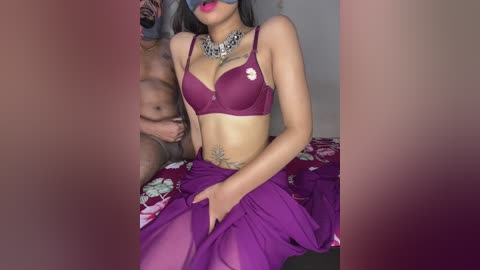 Video of a dark-skinned woman in a purple lace bra and sheer skirt, wearing a mask, sitting on a bed with a floral patterned sheet.