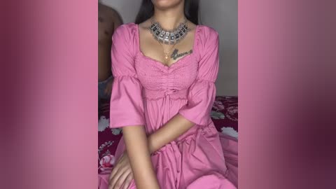 Video of a woman with long dark hair, wearing a pink ruffled dress with puffed sleeves and a statement necklace, sitting on a floral-patterned bed.