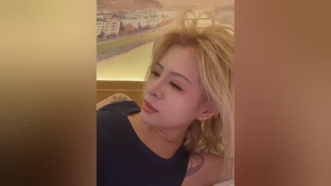 Video of a young woman with wavy blonde hair, wearing a navy blue top, leaning against a warm-toned wall. Background features a blurred mural of a cityscape.