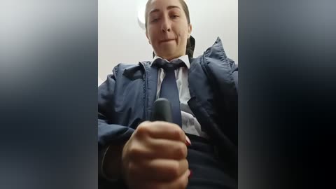 Video of a woman in a blue jacket, white shirt, and blue tie, holding a gray phone, taken from a low angle, partially obscured by a blurry background.