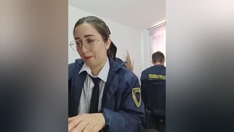 Video of a young woman with fair skin, brown hair tied back, wearing glasses and a navy police uniform, seated in a dimly lit room with two other officers.