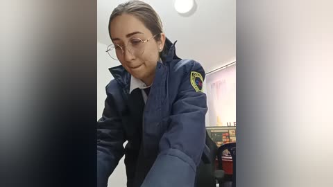 Video of a young woman with fair skin, brown hair in a ponytail, wearing glasses and a navy-blue police jacket with a badge, standing indoors with a blurred background.