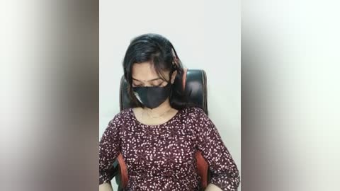 Video of a woman with medium brown skin, wearing a black face mask, dark hair, and a maroon floral-patterned blouse, seated on a black office chair against a plain white wall.
