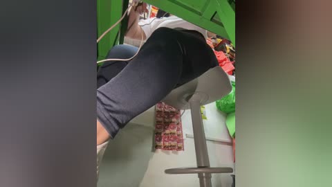 Video of a woman's lower body in black leggings and white shoes, bending over a green stool in a cluttered room with a red-patterned rug and assorted objects.