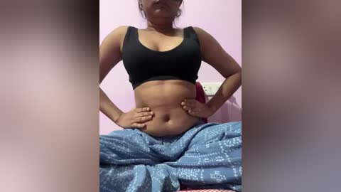 Video of a dark-skinned woman with medium-sized breasts wearing a black sports bra and blue patterned pants, seated on a bed with a light pink wall background.