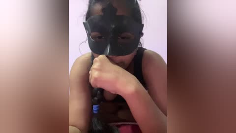Video of a woman with medium skin tone, wearing a black mask and black tank top, holding a black braided rope, against a blurry pink background.