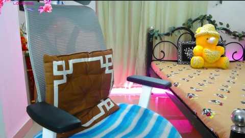 Video of a cozy, decorated room with a grey mesh chair, a brown cushion with white geometric design, a blue and white striped pillow, and a yellow teddy bear on a floral-patterned table.