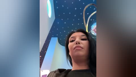 Video of a young woman with light skin, long black hair, and light makeup, standing in a colorful, futuristic room with a starry ceiling and metallic accents.