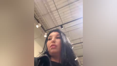 Video of a young woman with long black hair, fair skin, and full lips, looking down with a slight smile. She's in an industrial-style room with a high ceiling and exposed pipes.