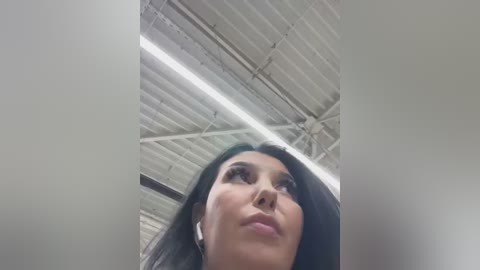 A low-angle video of a young woman with long, dark hair, wearing white headphones, looking upwards with a neutral expression. The background shows a white, corrugated metal ceiling with fluorescent lights.