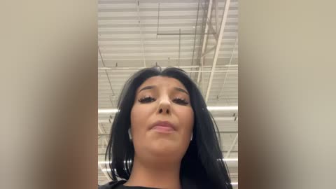 A video of a woman with long black hair and light skin, wearing heavy makeup including eyeliner and mascara, standing in an industrial warehouse with a high, ribbed ceiling.