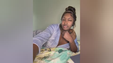Video of a young Black woman with medium brown skin, wearing a light blue robe, lying on a bed with a green and white patterned blanket. She has her hair in a messy bun and is smiling.