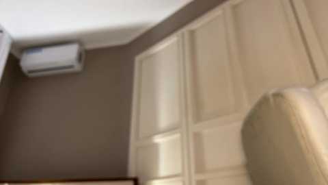 A video of a dimly lit room with beige walls and white paneling, featuring a mounted air conditioner on the left and a cushioned chair on the right.