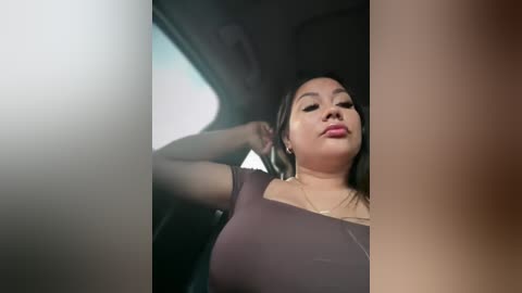 Video of a woman with long dark hair, light skin, and medium build, wearing a tight, brown top, sitting in a car. Her face is relaxed, eyes closed. The car's interior is visible, with a blurred window.