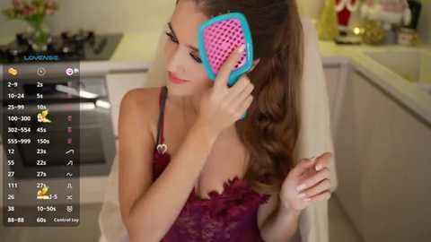 Video of a young woman with long brown hair, wearing a purple lace camisole, holding a blue and pink hairbrush, in a modern kitchen with white cabinets and a stove.