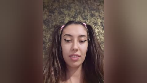 Video of a young Asian woman with long, wet brown hair styled in pigtails. She has light skin and wears a pink headband. Background is a textured, earthy-toned wall.