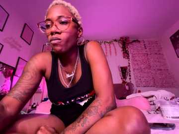 Video of a young black woman with short, bleached hair and multiple tattoos, wearing glasses, a black tank top, and a colorful belt, sitting on a bed in a dimly lit room with pink lighting.