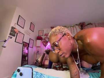 Video of a topless, slender, light-skinned woman with short, blonde dreadlocks and glasses, leaning on a bed in a dimly lit, cluttered room.