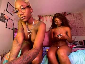 Video of two nude Black women with tattoos, one short-haired, one curly-haired, sitting on a bed in a cozy, colorful room with wall art and plants.