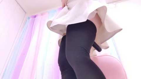 A video of a person's lower body, wearing black tights and a white, flowing dress, standing against a pastel-colored shower curtain. The image is bright and slightly blurry.