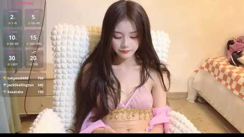 Video of an Asian woman with long black hair, wearing a pink bra and sheer pink robe, sitting on a bed in a cluttered room.