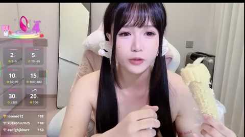 Video of a young Asian woman with long black hair, wearing a pink crown, sitting topless in a bedroom with a white bed and fluffy pillow, displaying a live stream overlay.