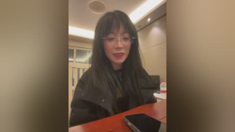 Video of an Asian woman with long black hair, wearing glasses and a black dress, seated at a wooden desk in a modern office.