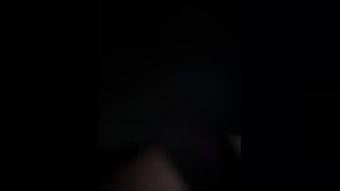 A dimly lit, grainy video of a person with short dark hair, wearing a dark jacket, standing in a shadowy, indistinct background. The image has a moody, mysterious atmosphere.