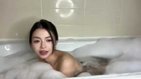 Video of an East Asian woman with long dark hair, partially submerged in a white bathtub filled with bubbles, giving a soft smile. Cream-colored tiled bathroom background.
