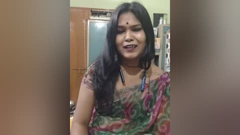 Video of a South Asian woman with long black hair, wearing a colorful saree with floral patterns, smiling in a cluttered room with wooden cabinets and clothes.