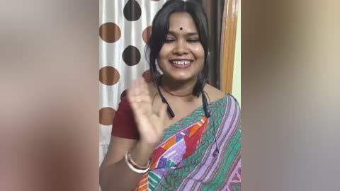 Video of a South Asian woman in traditional attire with a colorful sari, smiling and waving, against a background with a polka-dot curtain.