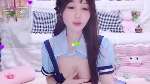 A video of an Asian woman with long, straight black hair, wearing a light blue maid outfit, lifting her top to reveal her breasts. Background features fluffy pillows, a teddy bear, and a houndstooth pillow.