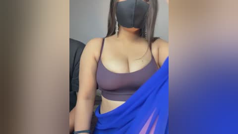 Video of a curvy, light-skinned woman with medium-length dark hair, wearing a purple sports bra, blue cloth, and black face mask. She's indoors, partially obscured by a beige wall.