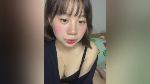 Video of an Asian woman with short brown hair and fair skin, wearing a black bra, blushing, in a bedroom with a colorful pillow.
