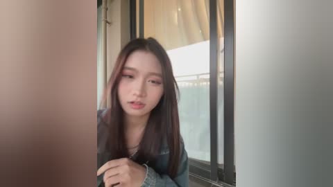 A video of an East Asian woman with long, straight, dark hair, wearing a denim jacket, standing in front of a window with light filtering in, looking slightly pensive.