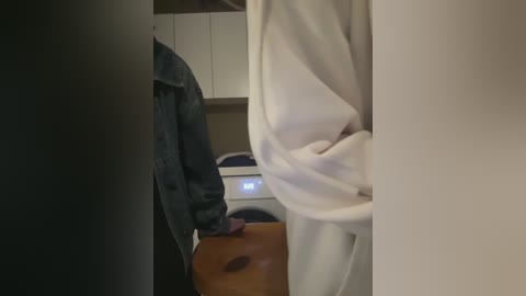 Video of a person in a white robe, standing in a laundry room with a dark denim jacket draped over their arm. A wooden laundry basket is on the floor, and a white washing machine is visible in the background.