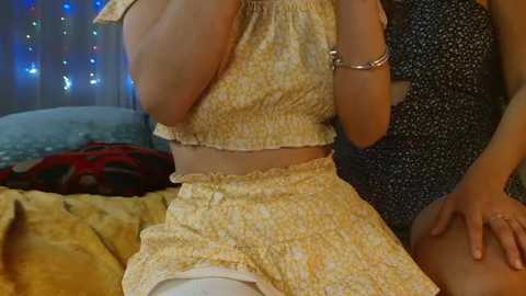 Video of a woman in a yellow floral crop top and matching skirt, holding a phone, with a person in a blue floral dress in the background, bed with blankets, and string lights.