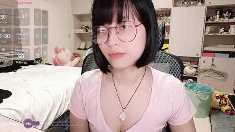 Media: Video of a young Asian woman with short black hair and glasses, wearing a pink top, sitting in a messy bedroom with a bed, toys, and a green bucket.