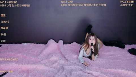 Video of an Asian woman with long brown hair lying on a pink fluffy blanket, wearing a light-colored top, with a microphone in her mouth, in a dimly lit room.
