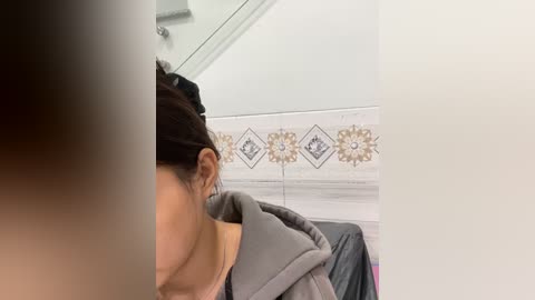 Media: Video of a woman with dark hair, wearing a gray sweater, seen from the side in a bathroom with white tiled walls and floral patterned accent tiles.