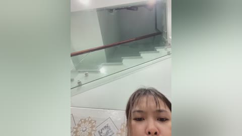 Video of an Asian woman with dark hair, light skin, and minimal makeup, standing at the bottom of a modern, glass-walled staircase in a bright, white room.