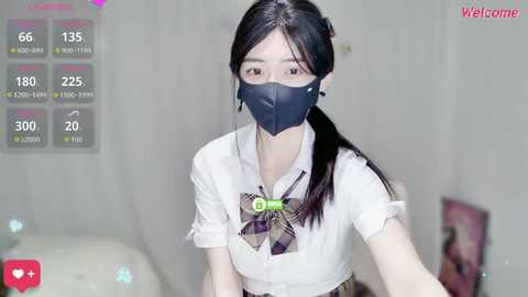 Media: A video of an Asian woman with long black hair, wearing a white blouse, plaid bow tie, and black face mask, standing indoors against a white backdrop.