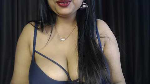 Video of a smiling, medium-skinned woman with long black hair, wearing a low-cut navy blue dress with a gold necklace, set against dark curtains.