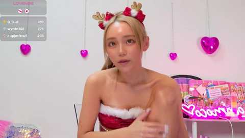Media: Video of a young blonde woman in a red and white Santa dress, with red and gold headband, in a pink-themed room with heart decorations and a \"Valentine's Day\" sign.