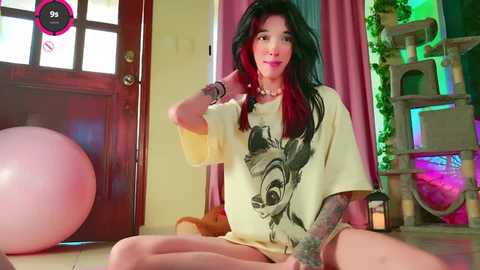 Media: Video of a tattooed woman with long black hair and red streaks, wearing a yellow graphic t-shirt, sitting cross-legged on the floor, surrounded by cat toys and a pink exercise ball.