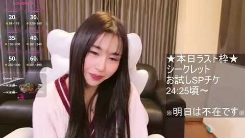 A video of an East Asian woman with long black hair, wearing a white robe, sitting on a black leather couch in a modern living room with beige curtains.