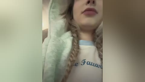 A close-up video of a woman with fair skin, light blonde hair in braids, wearing a white t-shirt with blue text, lying on a soft, green and white blanket.