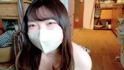 Media: Video of a young East Asian woman with straight, shoulder-length brown hair, wearing a white face mask and a white top, indoors, near a wooden shelf and a woven basket.