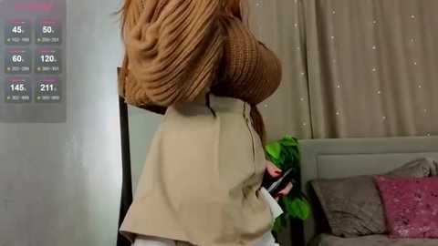 Media: Video of a woman with large breasts wearing a brown, ribbed sweater and beige skirt, standing in a bedroom with a white bed, pink pillows, and beige curtains.